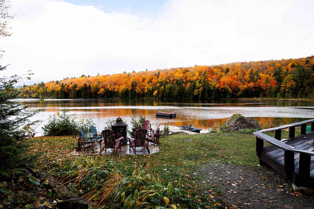 A scenic lakefront view features a circular firepit area surrounded by several seating options, set against a backdrop of vibrant autumn foliage reflecting in the calm water. A dock extends into the lake, with nature-inspired decorations visible along the shore.