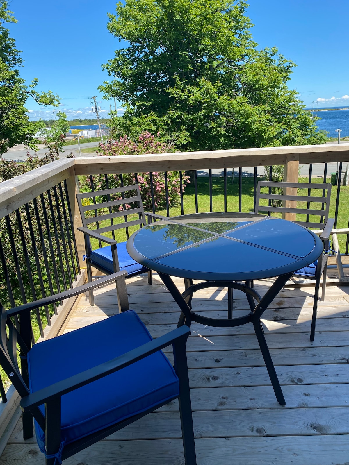 A private deck is shown featuring a round glass-top table surrounded by four black metal chairs with blue cushions. Lush greenery and the ocean are visible in the background under a clear blue sky.