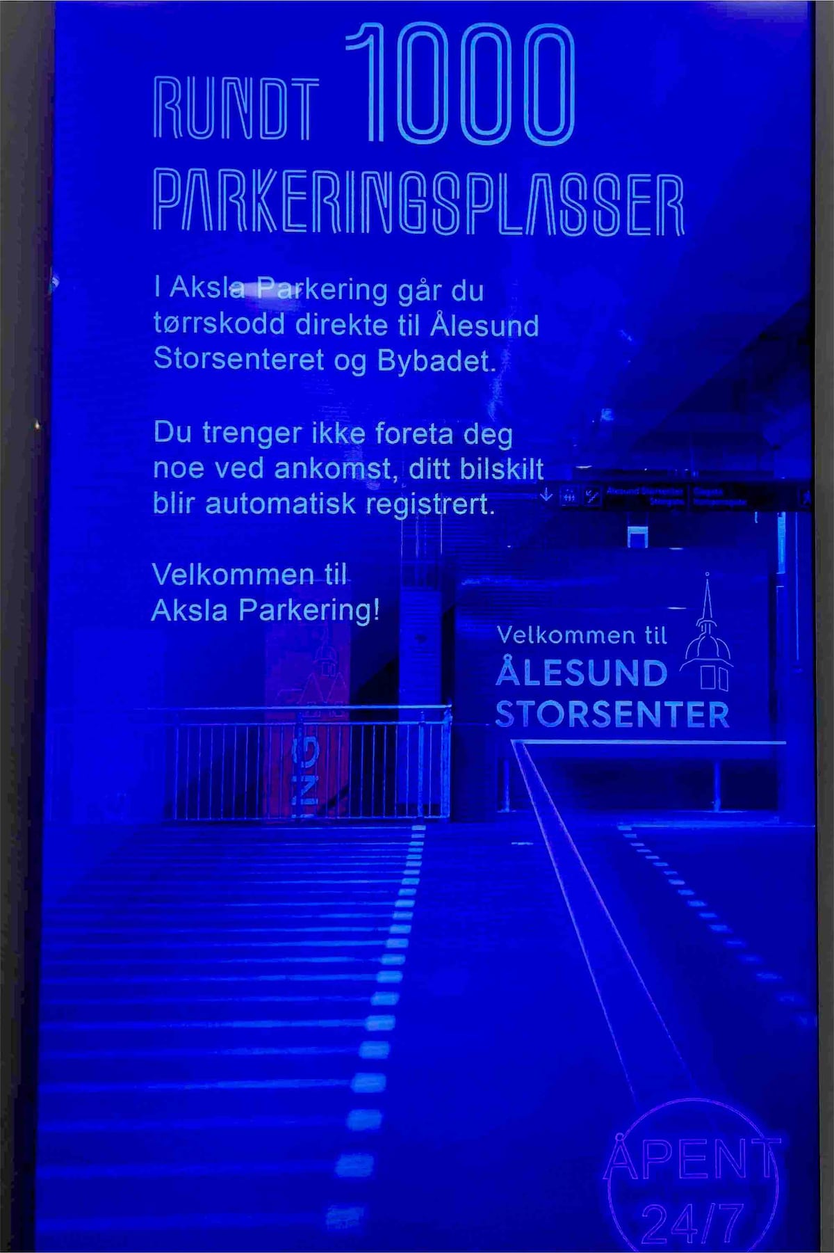 A bright blue informational sign displays details about the Aksla Parking facility. The text indicates the availability of approximately 1,000 parking spots, with instructions for automated registration upon arrival. The modern design emphasizes clear, easy-to-read lettering against the vibrant background.