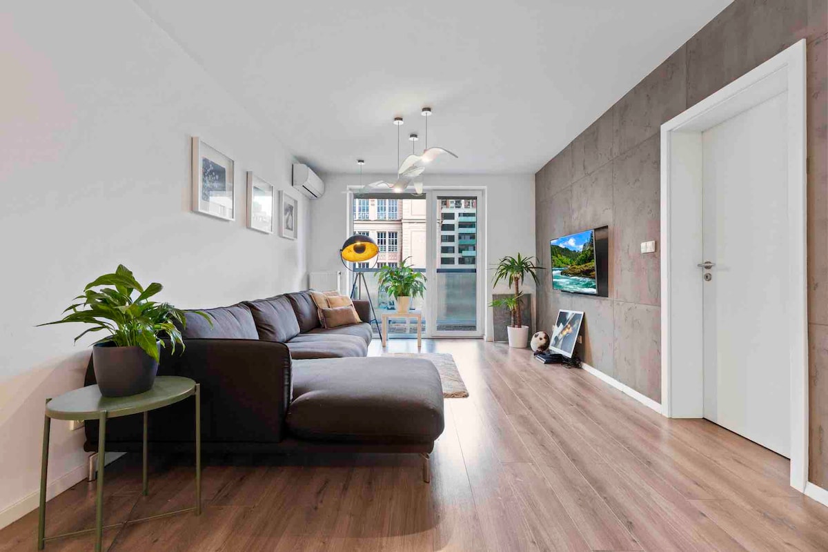 A spacious living area features a large gray couch and a round side table with a potted plant. Natural light fills the room through glass doors leading to a balcony. A TV is mounted on the wall, and artwork is displayed on the adjacent wall.