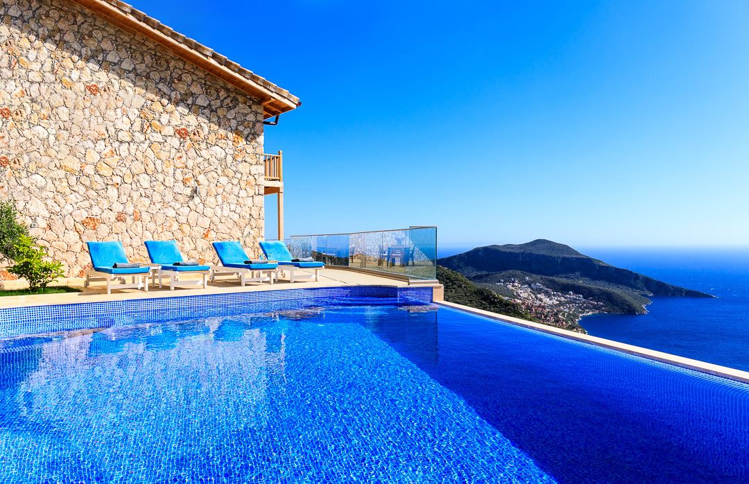 An infinity pool is visible, featuring clear blue water that mirrors the sky. Surrounding the pool, several lounge chairs are arranged, offering a view of the coastal landscape and mountains in the distance. A stone exterior building adds texture to the serene environment.