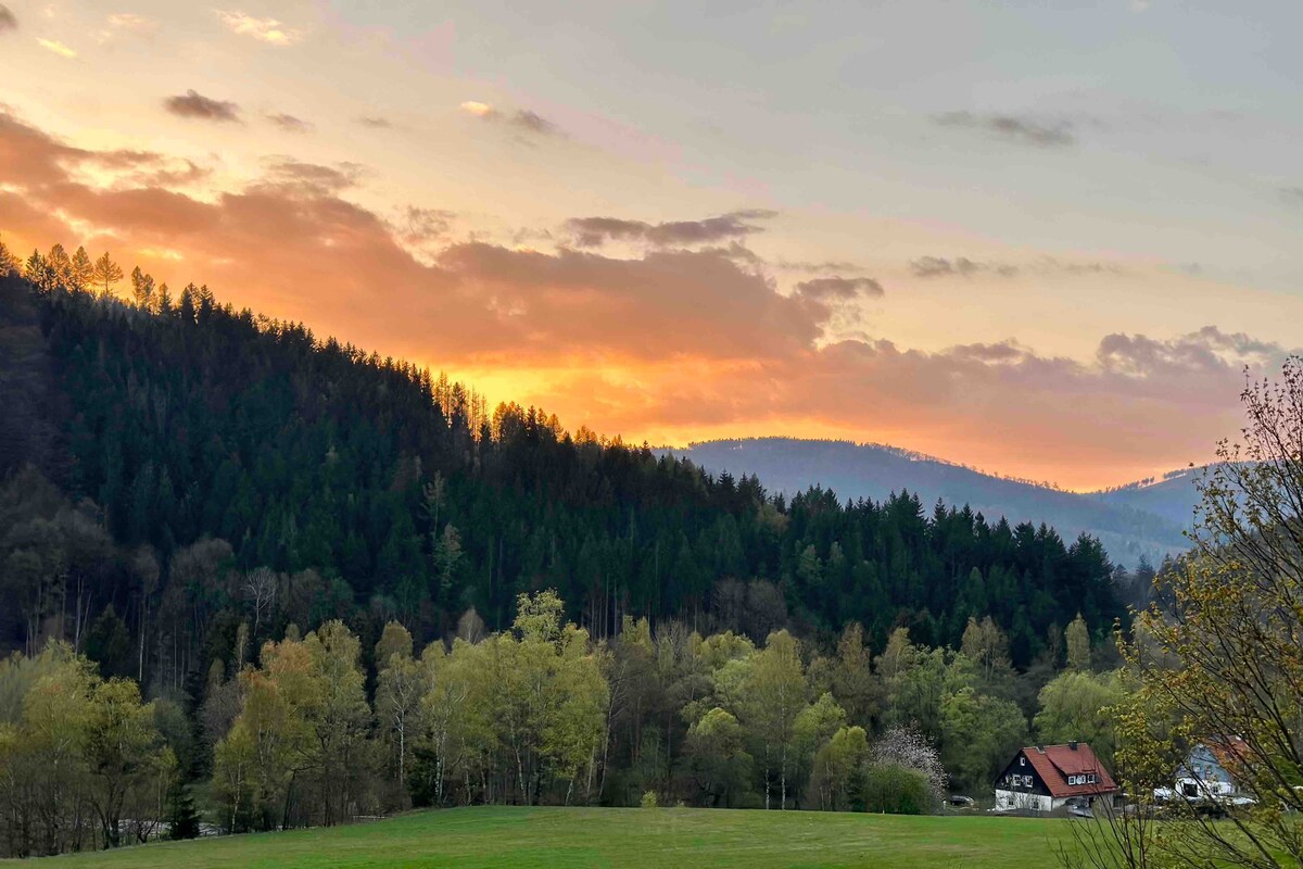 A serene landscape is depicted, featuring rolling hills covered with trees under a colorful sunset. The sky is filled with soft hues of orange and pink, blending into the twilight. A distant house is visible amidst the greenery, set against a backdrop of mountains.