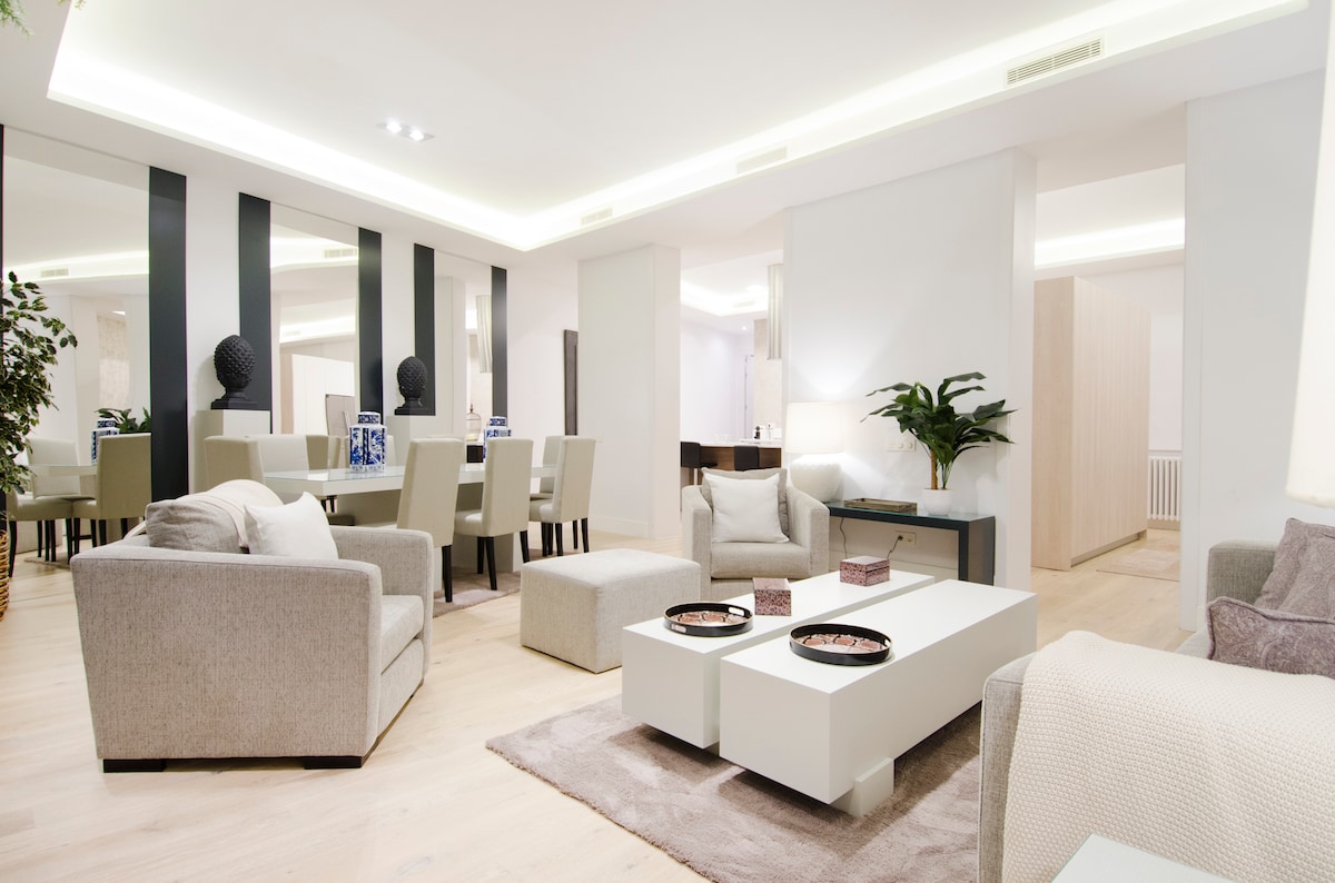 A modern living area showcases an open-plan design featuring soft seating and a coffee table. Light wood flooring complements the space, while large windows allow natural light to fill the room. A dining area with a large table is visible in the background.