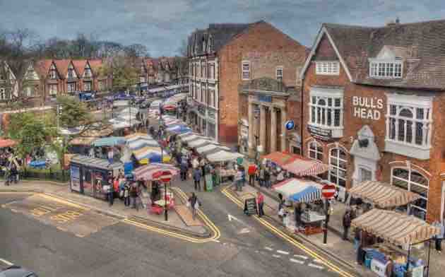 A bustling street market is captured from above, featuring various colorful stalls and awnings. Nearby, a brick building with the sign 'BULL'S HEAD' adds character to the scene. Visitors can be seen exploring the market, surrounded by trees and cheerful vendors.