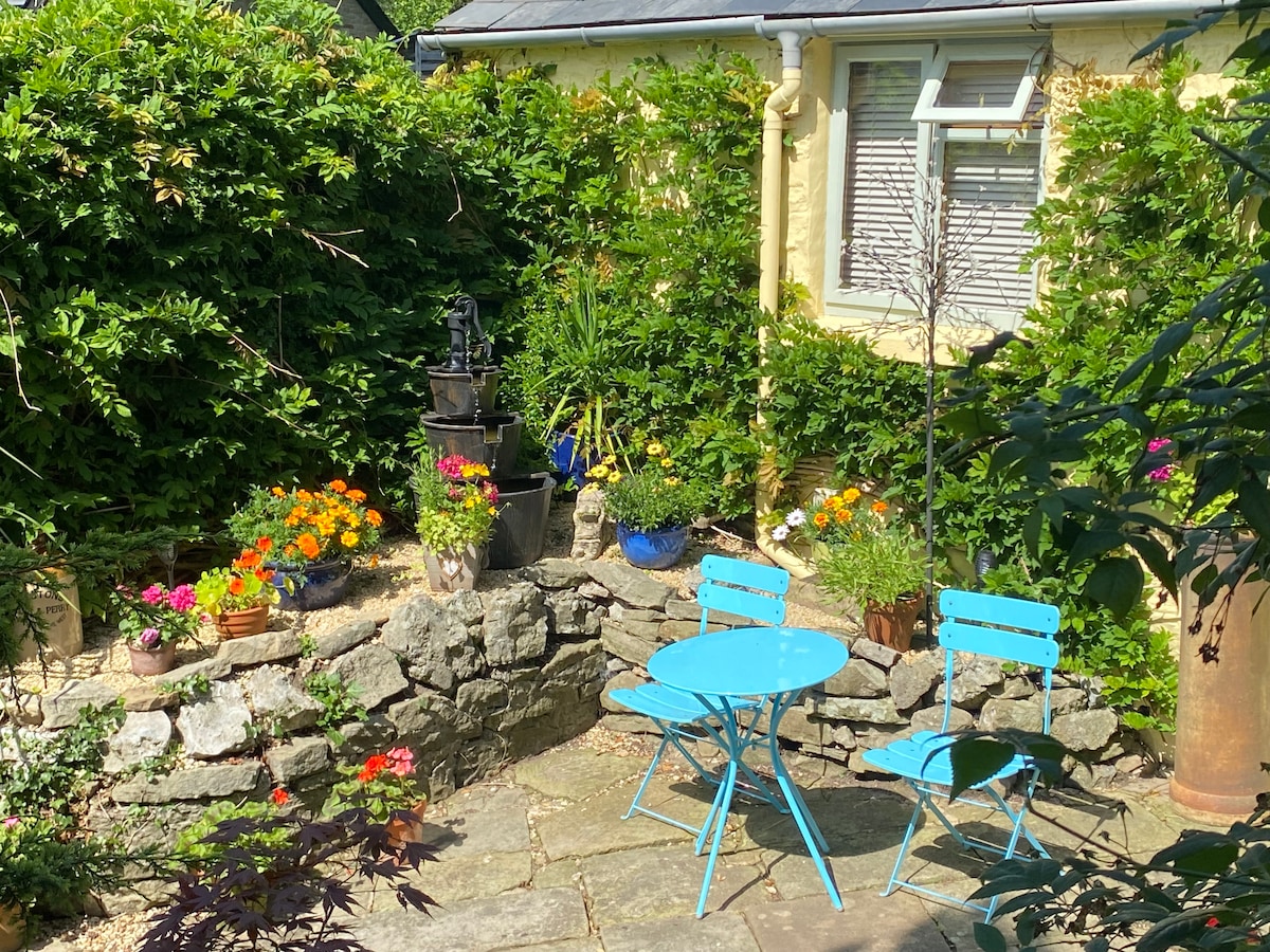 A charming outdoor space features a stone wall surrounded by vibrant flower pots and green foliage. Blue bistro table and chairs are positioned on the stone patio, creating a peaceful setting. A water feature adds a soothing element to the serene garden atmosphere.
