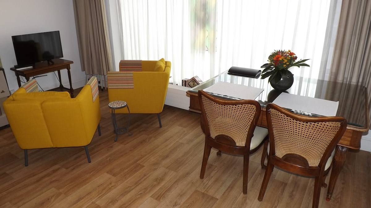 A cozy sitting area is highlighted by two yellow armchairs and a circular side table. A wooden dining table adorned with a floral centerpiece is accompanied by two woven chairs. Floor-to-ceiling windows allow natural light to fill the space, creating a bright and welcoming atmosphere.