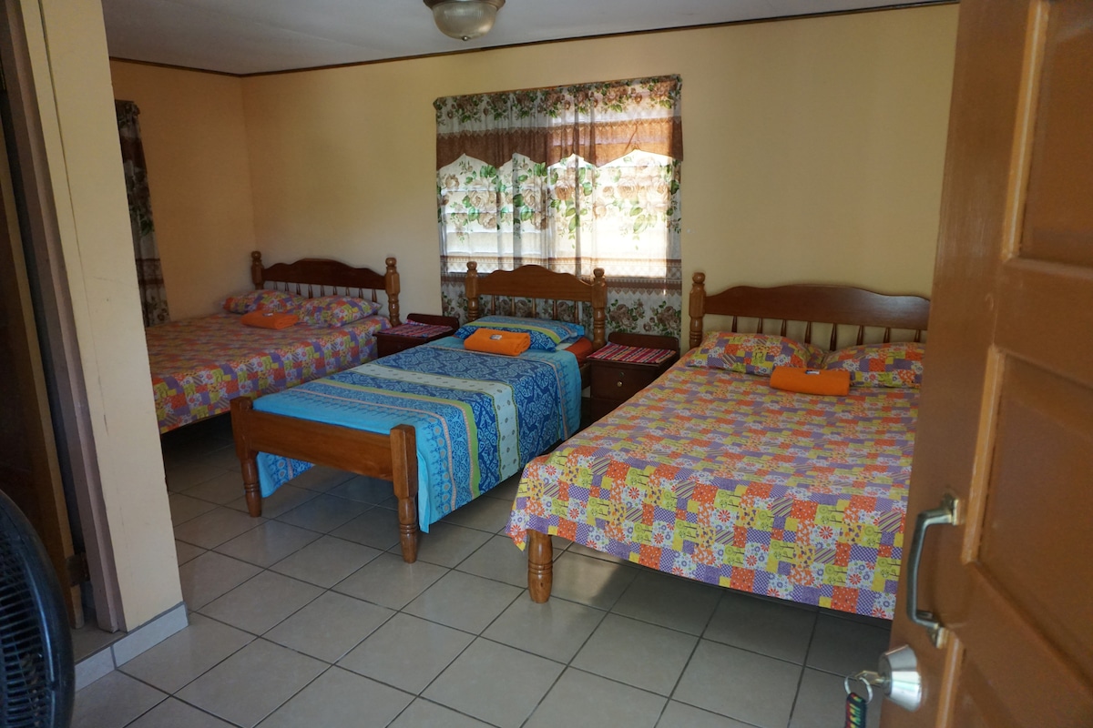 The sleeping area contains three spacious beds arranged with colorful patterned linens. Bright orange towels are neatly placed on each bed. A large window, adorned with lace curtains, allows natural light to illuminate the room. Tiled flooring contributes to the simple, clean aesthetic.