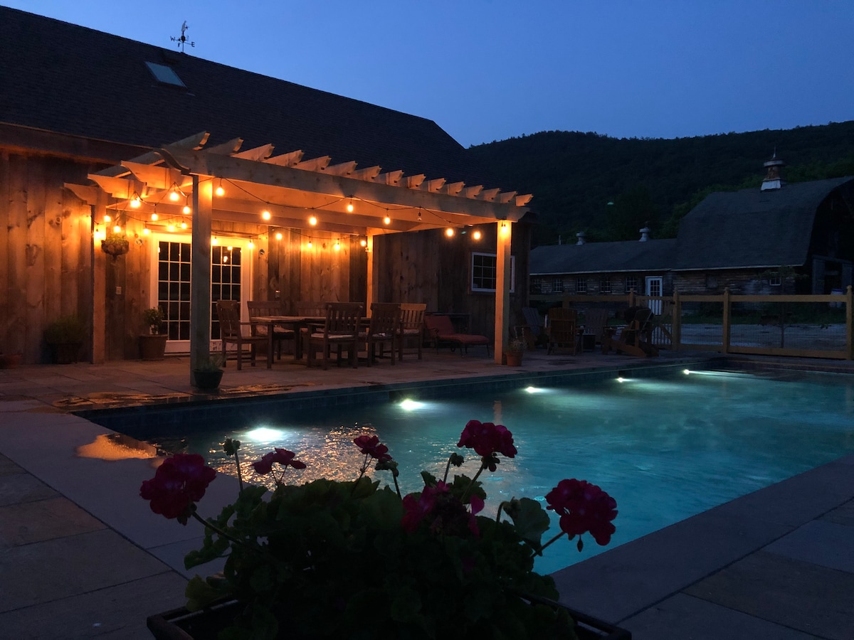 An inviting outdoor space is illuminated by warm string lights, showcasing a pool with softly glowing lights. A pergola stands over a dining area surrounded by wooden furniture, while vibrant flowers add a pop of color to the scene.