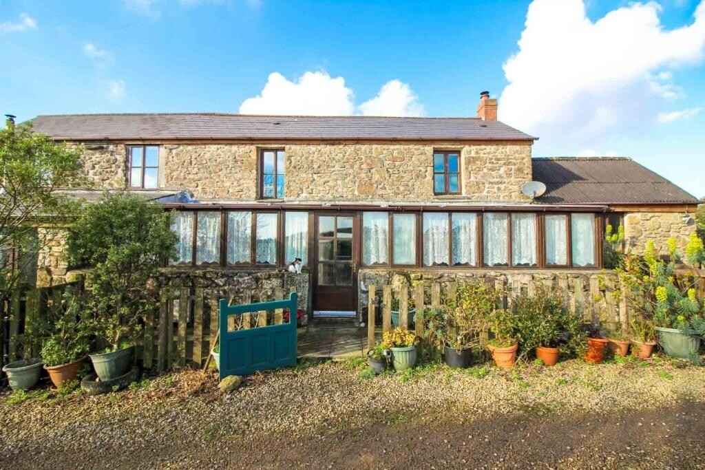 The exterior of the cottage is presented, featuring a charming stone facade and a welcoming entrance. Large windows allow natural light to fill the interior. Surrounding greenery and potted plants enhance the rural setting, with a peaceful lane leading up to the door.