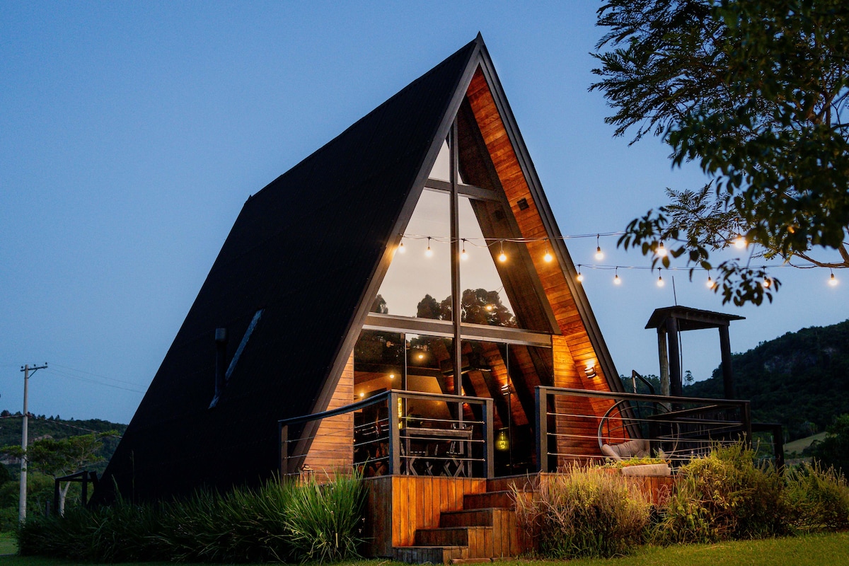 The cabin features a distinctive A-frame structure with large glass windows, allowing for ample natural light. The exterior is complemented by wooden accents and a welcoming entryway, surrounded by lush greenery and ambient outdoor lighting.