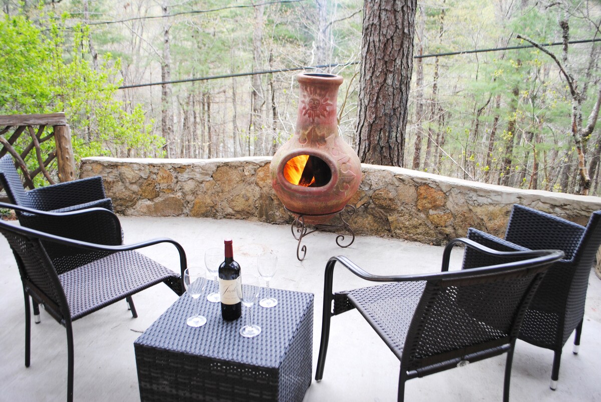 An outdoor seating area features four woven chairs arranged around a small table. A bottle of red wine and two glasses are placed on the table. A clay chiminea stands nearby, glowing with fire, surrounded by trees in a tranquil natural setting.