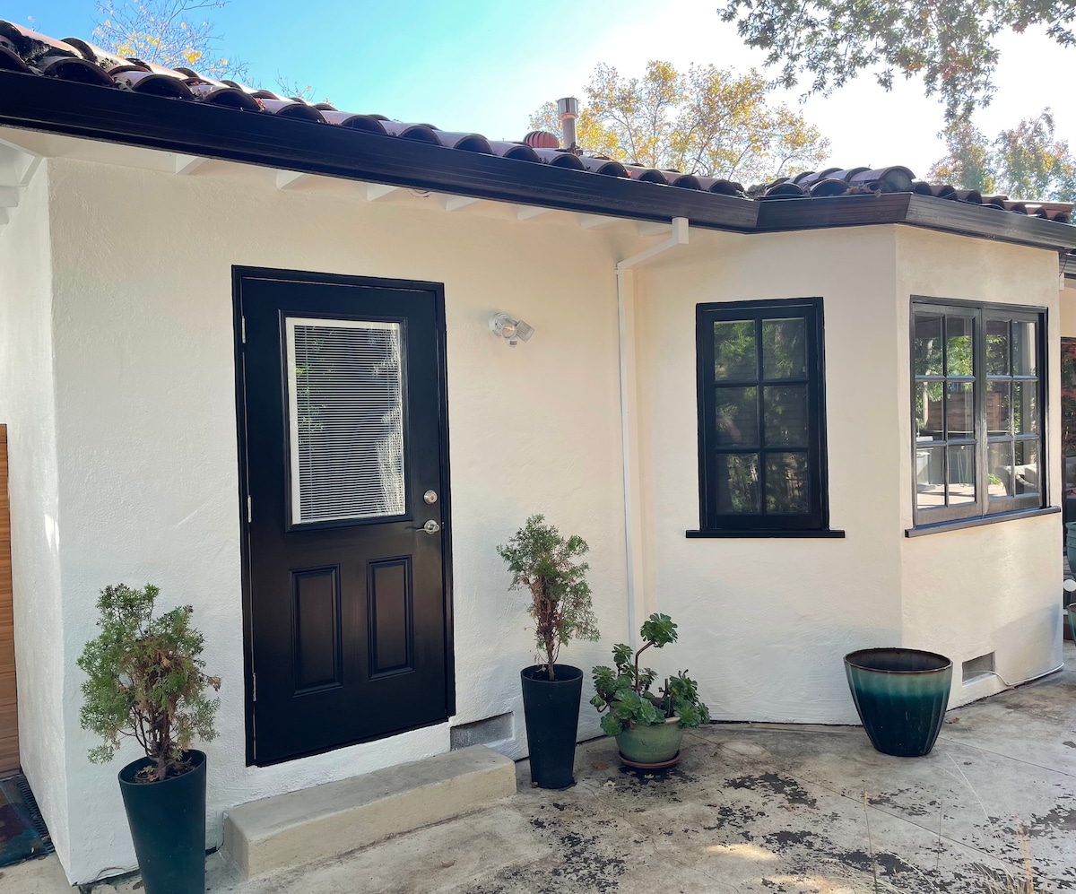 A private entrance to the suite is framed by a white exterior wall, featuring a black door and a small window. Potted plants in varying sizes decorate the entryway. Natural light brightens the space, with a clear blue sky visible above.