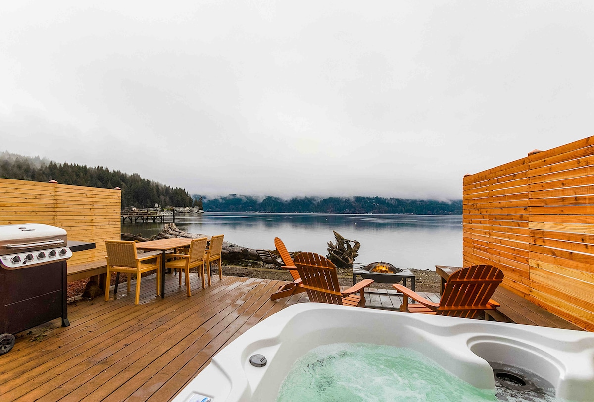 A spacious deck offers a hot tub overlooking the water and a firepit surrounded by wooden seating. A dining table stands ready for outdoor meals, all set against a backdrop of serene water and misty hills.
