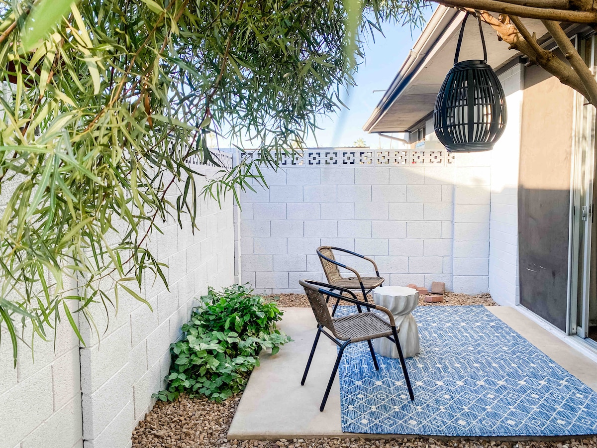 A private outdoor patio space features two woven chairs positioned around a small table. A patterned blue rug adds comfort beneath them. The area is shaded by overhanging greenery and surrounded by a white block wall, providing a serene nook for relaxation.