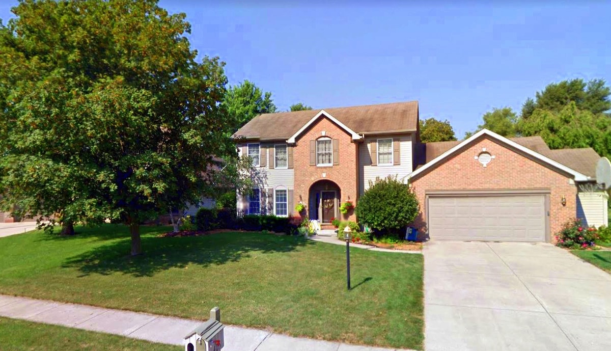 The exterior of a two-story brick house is presented, featuring a well-maintained lawn and a concrete driveway. Lush trees and vibrant shrubbery frame the entrance, which includes a covered porch and stylish entryway with decorative elements. A mailbox is visible at the street.