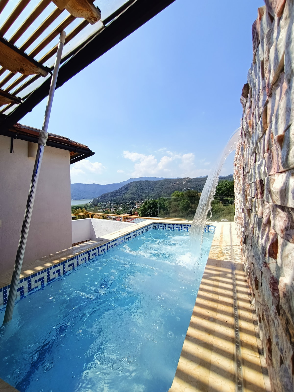 A serene swimming pool is featured, with water cascading elegantly from a stone wall. The mountain backdrop provides a scenic view under a clear blue sky, while sunlight filters through a wooden pergola, creating a tranquil outdoor space.
