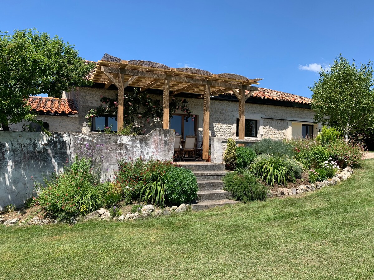 The exterior view of Gite L'Ecurie features a charming entrance with wooden pergola, flanked by vibrant plants and neatly arranged flowers. Stone steps lead up to large glass doors, revealing the interior. Green lawns extend beyond the landscaped garden, enhancing the natural setting.