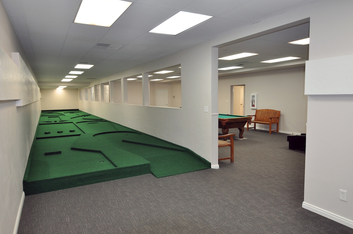 An indoor recreation area features a long, green carpet mimicking a miniature golf course. Bright overhead lights illuminate the space, which includes several openings along the walls providing views into adjacent areas. A wooden bench and a billiard table are visible in the background.