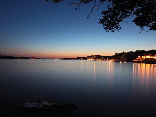 A serene lakeside view at twilight captures still waters reflecting soft hues of blue and orange. Gentle lights dot the shoreline, suggesting nearby homes and activity. The image is framed by the silhouette of trees, enhancing the peaceful atmosphere of the setting.