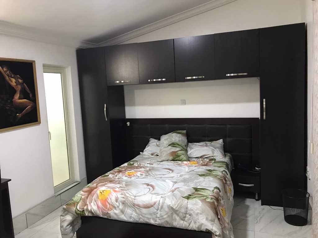 A well-furnished bedroom features a spacious bed with a floral bedding set. A dark wood wardrobe is positioned above a headboard, providing ample storage. Natural light is filtered in through a sliding glass door, which leads to a small balcony.
