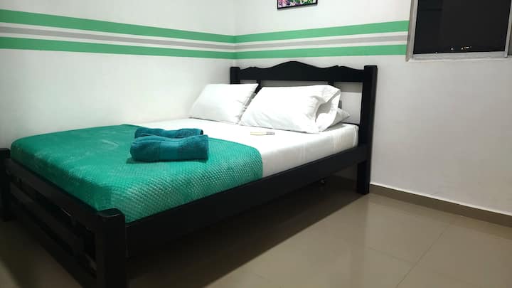 Chumba cha watu wawili kilicho na feni ya sakafu na dirisha kubwa ambalo upepo wa bahari unaingia.
