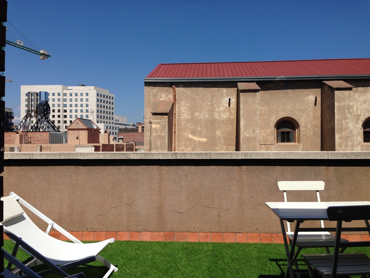 A private terrace features modern outdoor furniture, including white chairs and a small table set against a backdrop of a clear blue sky. The terrace has artificial grass, and the view includes surrounding buildings with subtle architectural details.