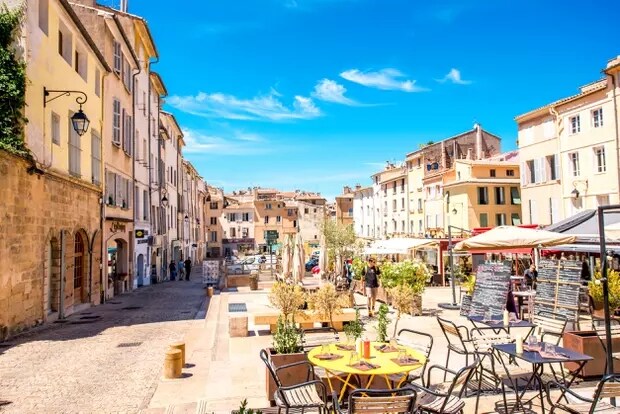 A charming street scene is depicted with cafe tables and umbrellas set along a cobblestone path. Colorful buildings line the street under a clear blue sky. People can be seen leisurely exploring the area, with lush greenery and decorative planters adding a touch of nature.
