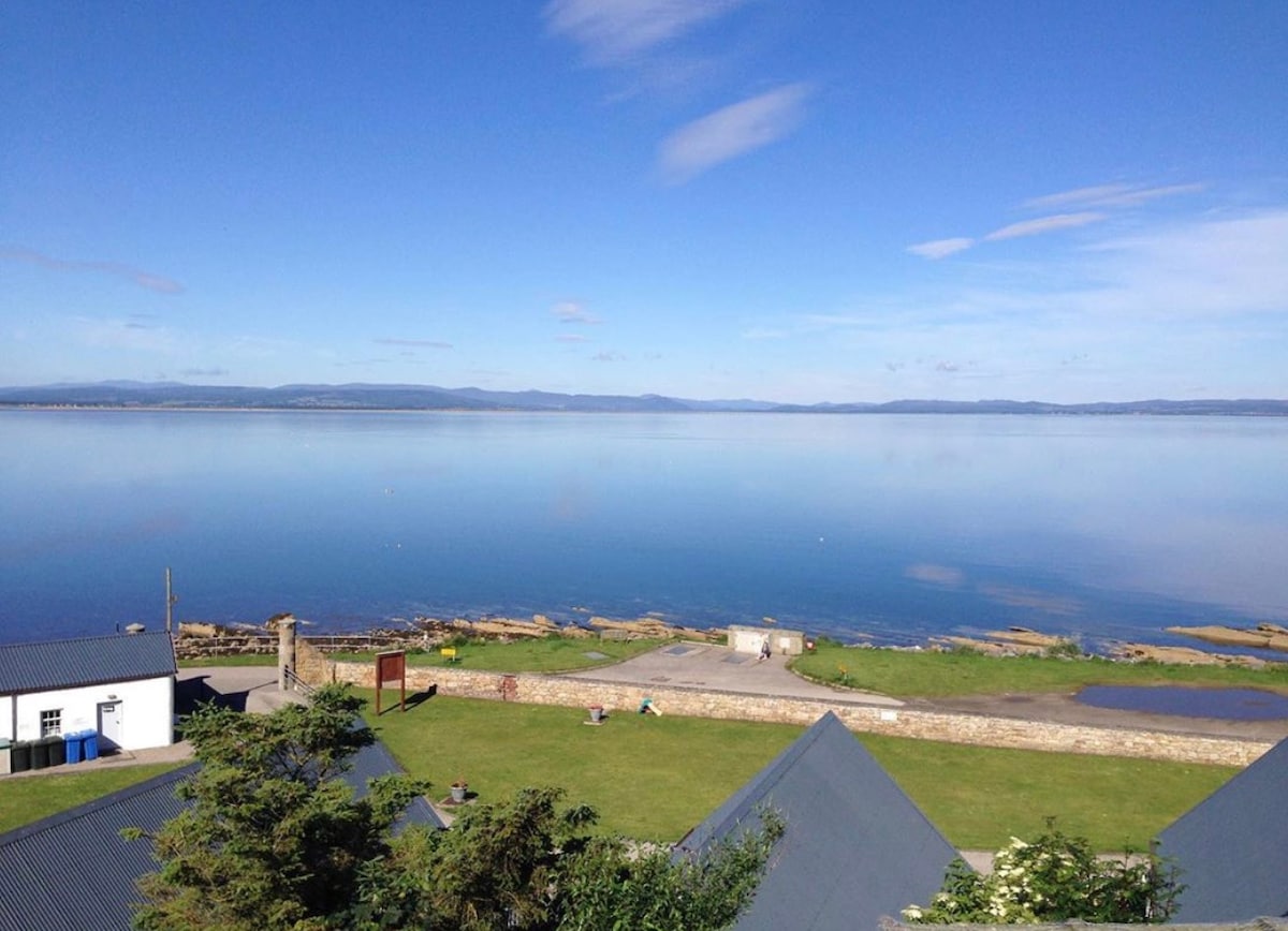 A panoramic view captures the calm waters of the Dornoch Firth, reflecting a clear blue sky. The green landscape extends along the shoreline, with well-maintained grassy areas visible, enhancing the coastal ambiance. Nearby rooftops are seen dotting the foreground.