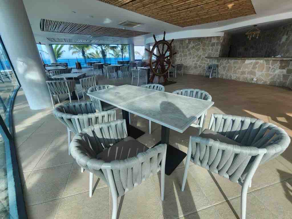 An elegantly arranged dining area features a central table surrounded by six uniquely designed chairs. Natural light pours in through large windows, highlighting the textured stone wall and wooden ceiling. A decorative ship's wheel adds a nautical element to the inviting space.