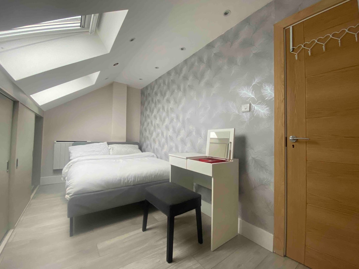 The loft space features a spacious bed with white linens, accompanied by a mirror on a minimalist desk. Natural light floods the room through a skylight, highlighting the textured wall design and the sleek wooden door.