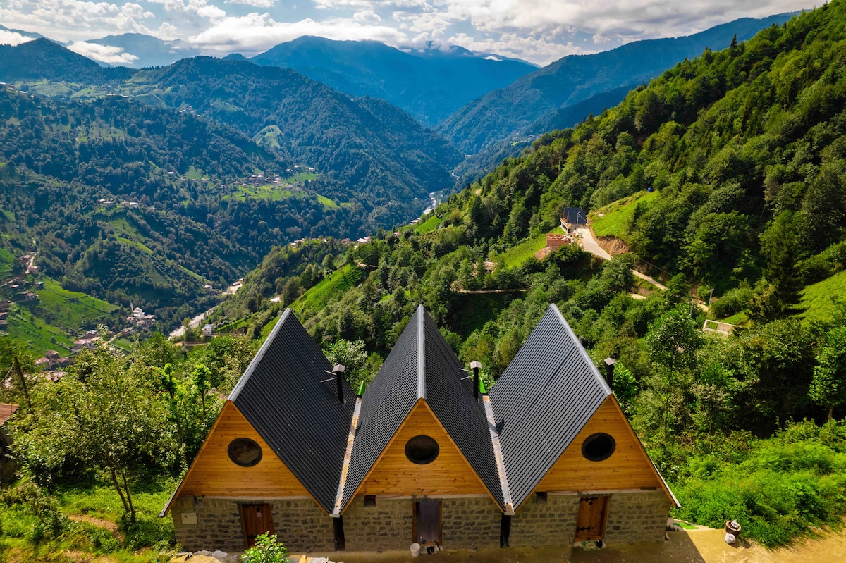 Three charming cabins are situated on a hillside, surrounded by lush green mountains. Their triangular roofs contrast with the blue sky and rolling hills in the background, creating a serene view of the valley below.