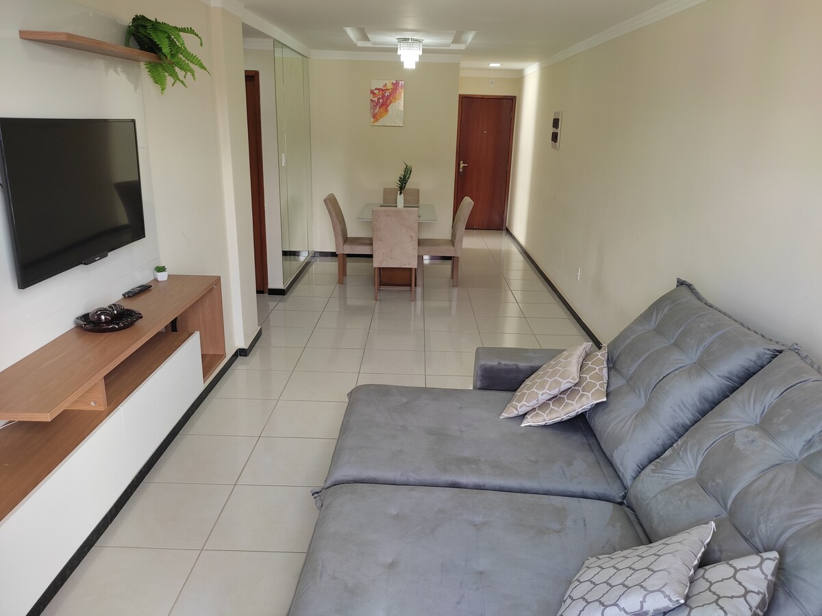 A spacious living area features a grey sofa with decorative cushions, positioned near a wall-mounted television. A dining table with four chairs is visible in the background, complemented by a modern light fixture overhead. The room is well-lit, showcasing neutral wall tones and tiled flooring.