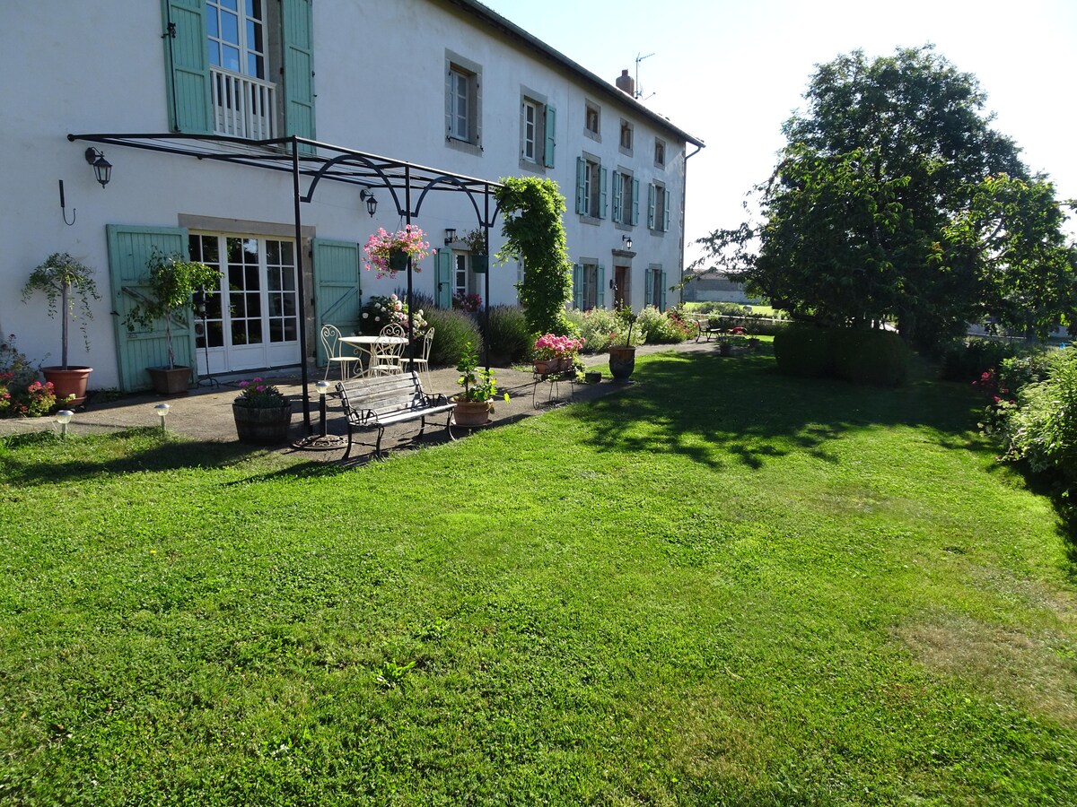 A large grassy area surrounds the entrance of the gîte, framed by various plants and flowers. A seating area with metal chairs and a table is positioned under a decorative pergola, enhancing the outdoor space. Light streams down from a clear blue sky.