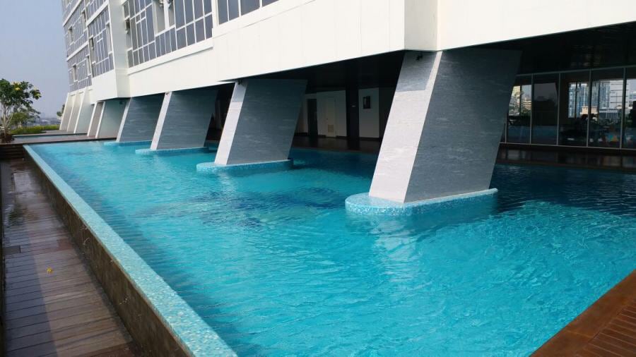 A swimming pool is viewed from an elevated position, featuring clear turquoise water and smooth, rectangular pillars supporting the structure above. The surrounding wooden deck enhances the pool area’s spacious feel, while large windows can be seen reflecting the exterior landscape.