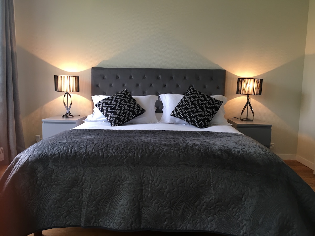 A spacious bed is presented with a luxurious grey coverlet and decorative pillows. Soft bedside lamps provide gentle illumination on either side of the bed, and textured wall panels add an elegant touch to the room's decor.