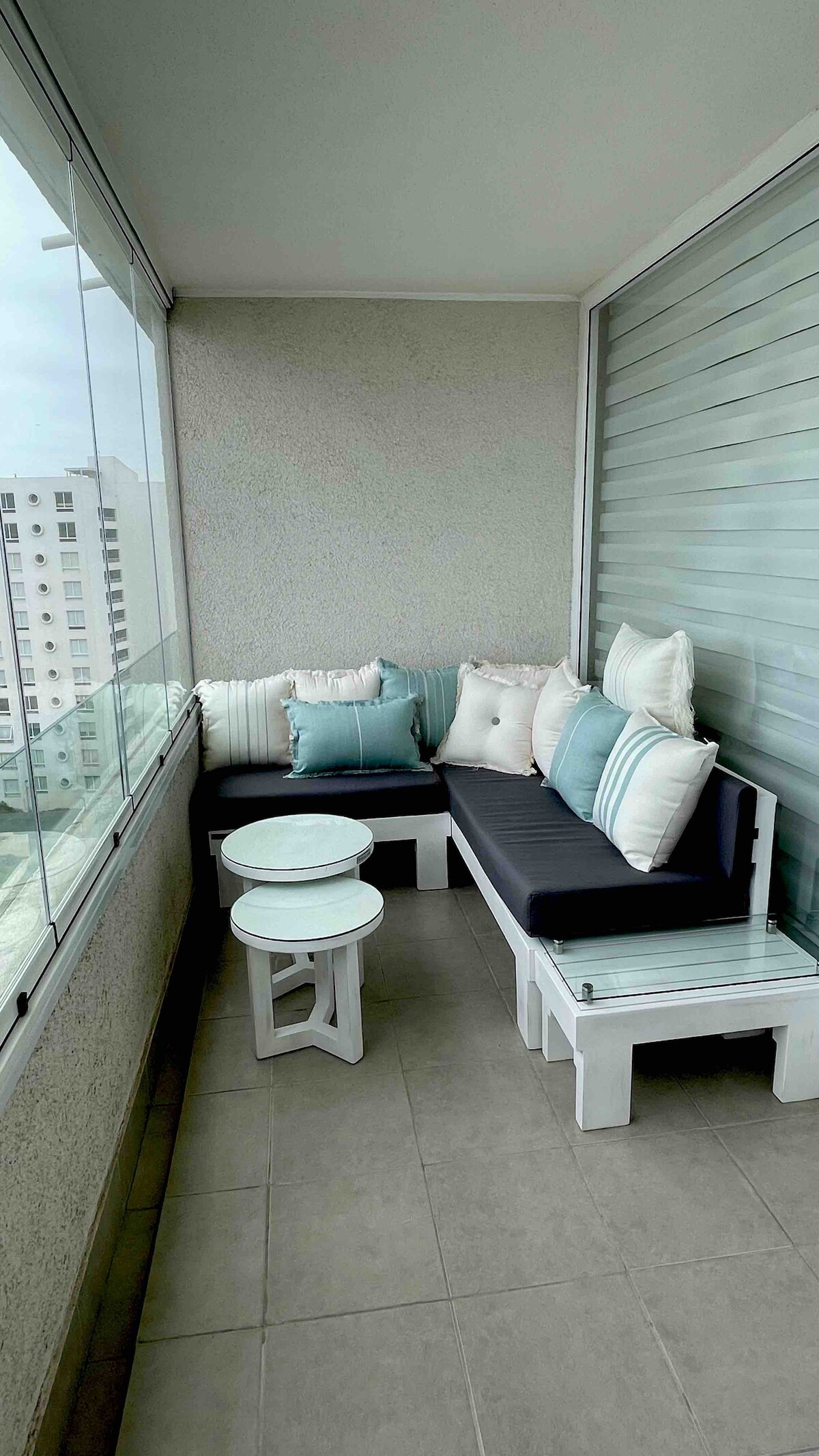 A cozy balcony seating area is showcased, featuring a black cushioned L-shaped sofa adorned with white and teal pillows. A small round table is placed in the center, and floor-to-ceiling windows provide a view of the surrounding cityscape.