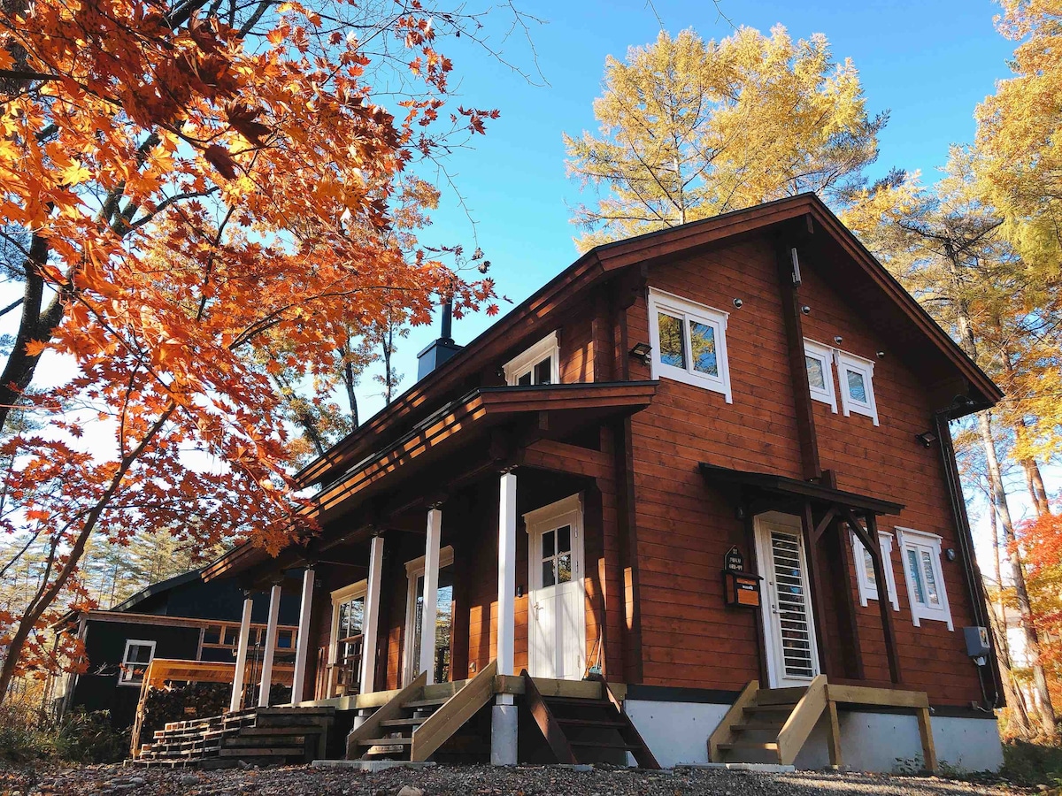 The lodge's wooden exterior is framed by vibrant autumn foliage, showcasing a welcoming entrance with a covered porch and steps leading up to the door. The structure features multiple windows that enhance natural light, set against a clear blue sky.