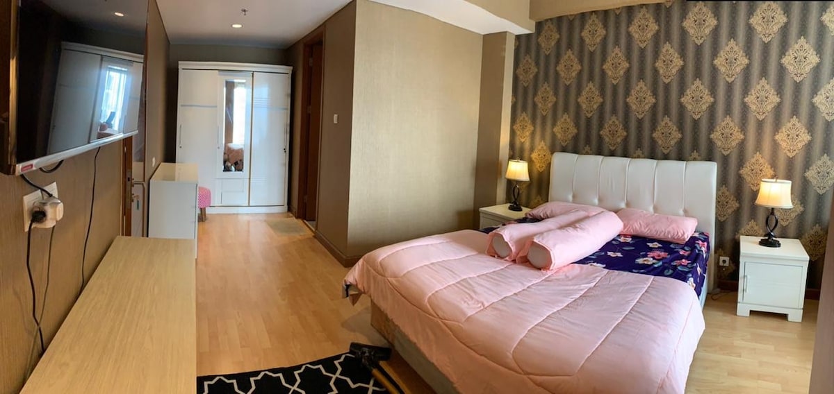 A spacious bedroom features light wood flooring and soft pink bedding on a large bed. Two nightstands with lamps are positioned beside the bed. A large wardrobe is visible in the background, and a flat-screen television is mounted on the wall opposite the bed.