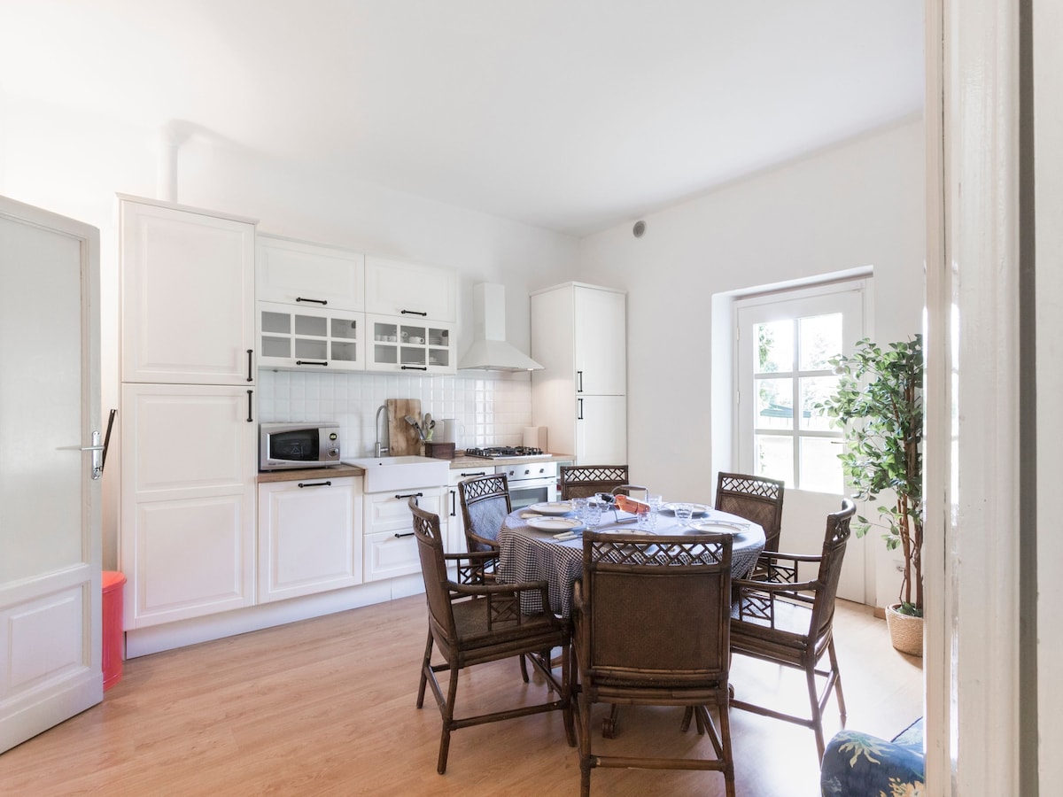 The kitchen features a round dining table surrounded by eight woven chairs. White cabinetry and appliances are complemented by a simple tile backsplash. Natural light enters through a large window, illuminating the space and enhancing the inviting atmosphere.