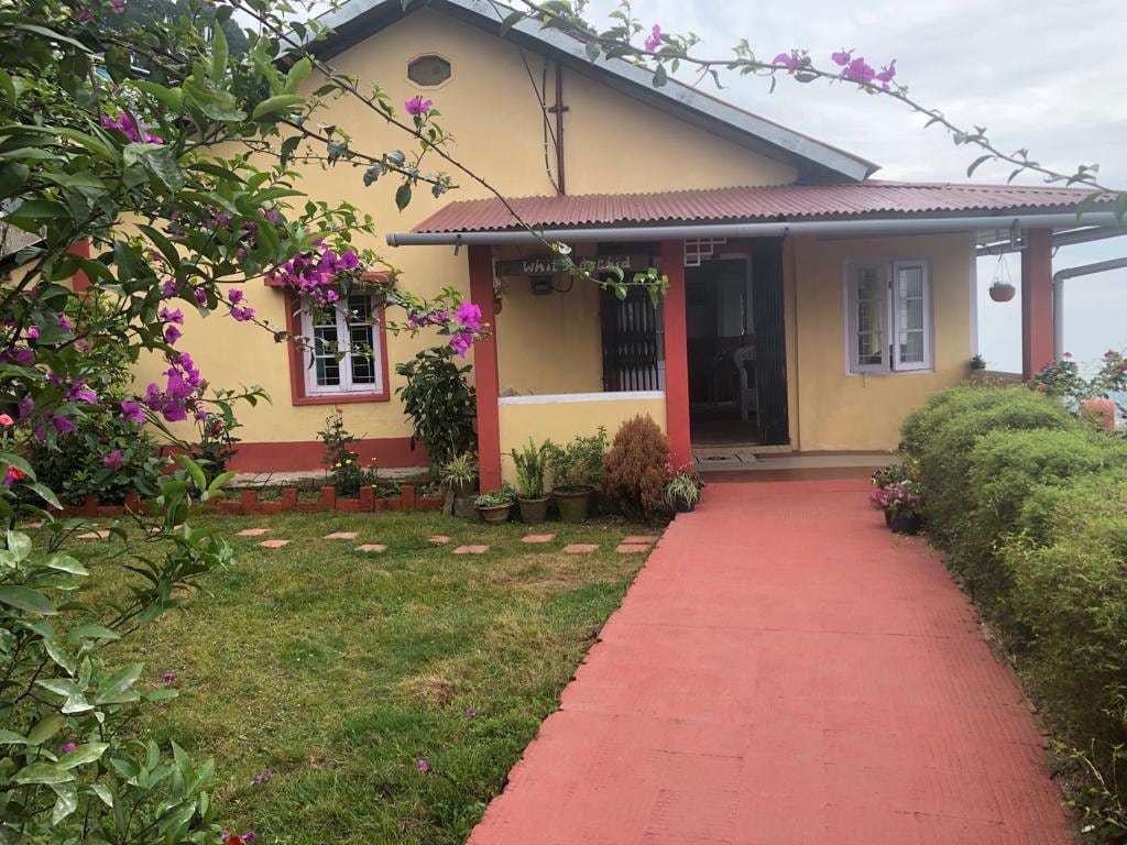 A charming guest house is set against a backdrop of lush greenery, with vibrant flowers framing the entrance. A red pathway leads to the inviting porch, where light-colored walls contrast with a dark roof. Windows are adorned with small potted plants, enhancing the welcoming ambiance.
