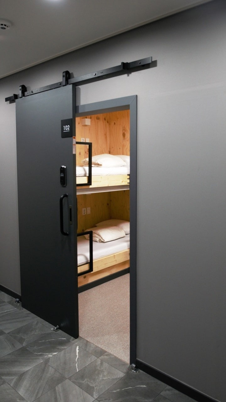 An open industrial-style sliding door reveals a capsule bed room. Wooden bunk beds are neatly arranged inside, with white bedding visible. The walls are painted in a soft grey, complementing the modern aesthetic and maintaining a clean and minimalist feel.