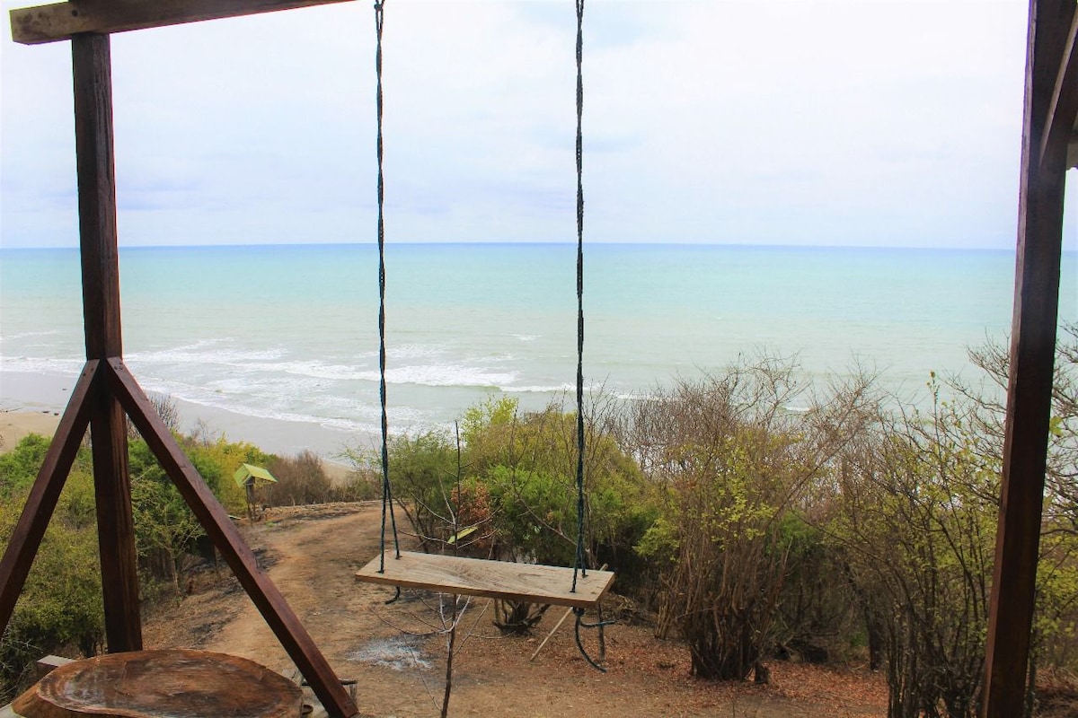 A wooden swing hangs from a sturdy structure, providing a clear view of the calm sea and distant shoreline. Lush greenery is visible in the foreground, with a sandy path leading toward the beach under a cloudy sky.
