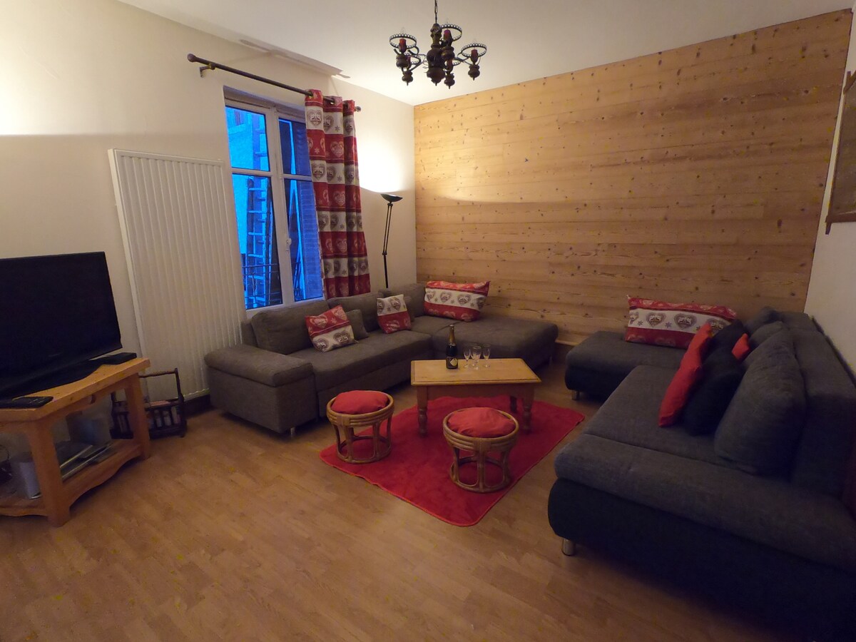A cozy living area features a mix of sectional sofas and a coffee table, all arranged on a wooden floor with a vibrant red area rug. Natural light enters through a large window, complemented by decorative curtains. A TV is positioned on an entertainment unit.