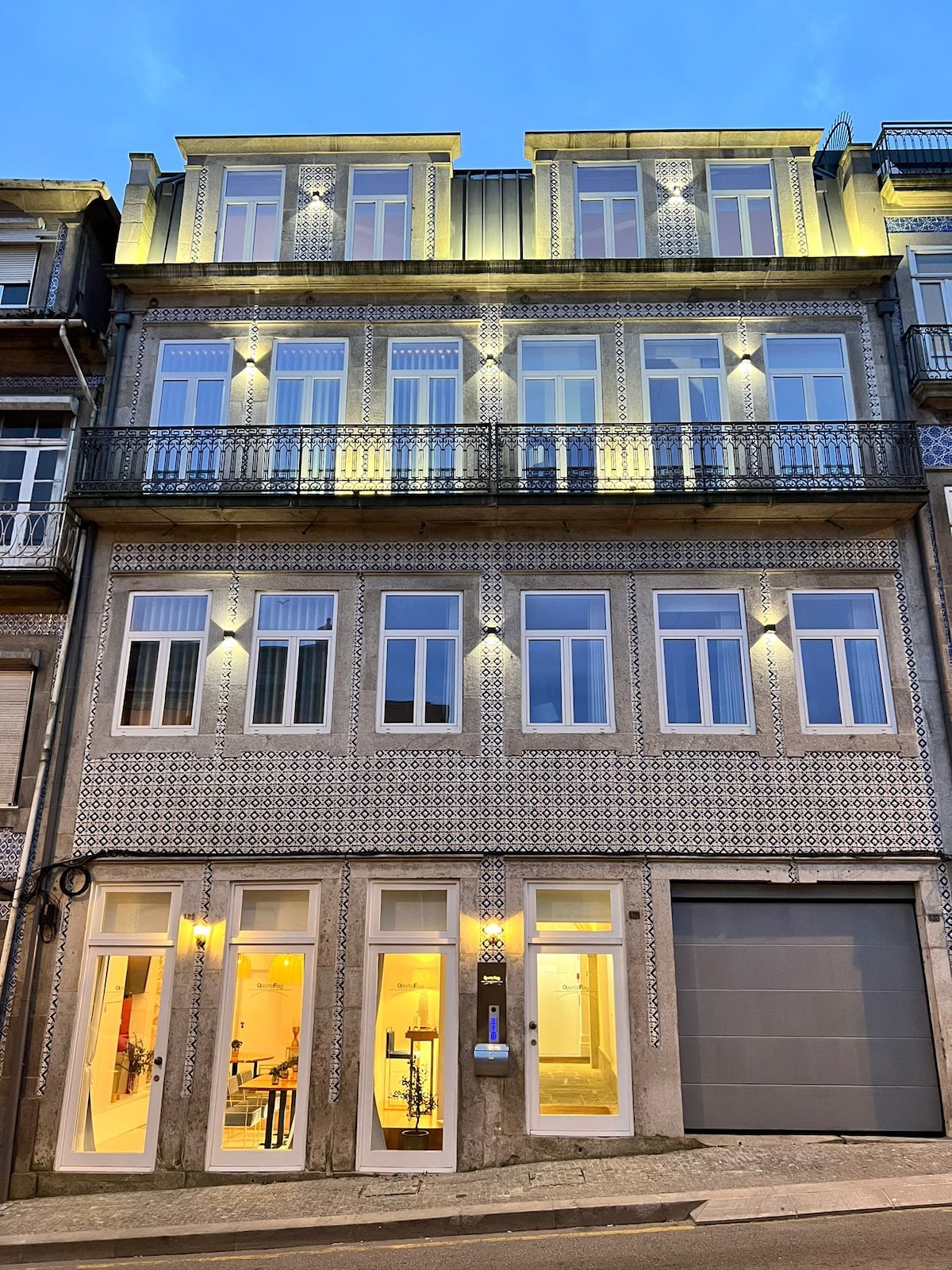 The exterior of a multi-story building is presented, featuring decorative tile work and illuminated windows. Soft yellow lights highlight the ground floor, where large glass doors provide a glimpse into the interior. A garage door is visible on the right side.