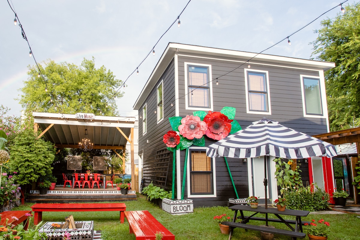 A charming two-story villa is presented, featuring oversized flower decorations at the entrance. Colorful outdoor seating areas are visible, adorned with striped umbrellas and vibrant red picnic tables. A cozy covered patio with a chandelier offers additional seating, creating an inviting atmosphere for relaxation.