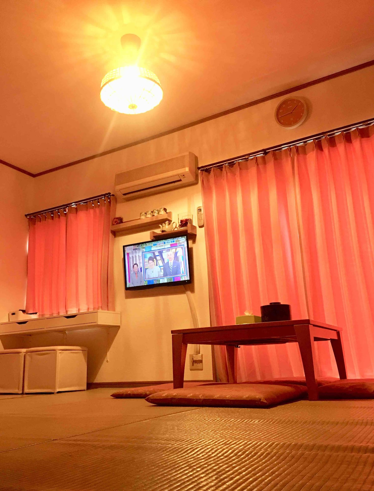 The room is warmly lit, featuring a low table in the center and tatami flooring. Two large, pink curtains frame a window, allowing soft light to filter in. A television is mounted on the wall opposite, providing entertainment for guests.
