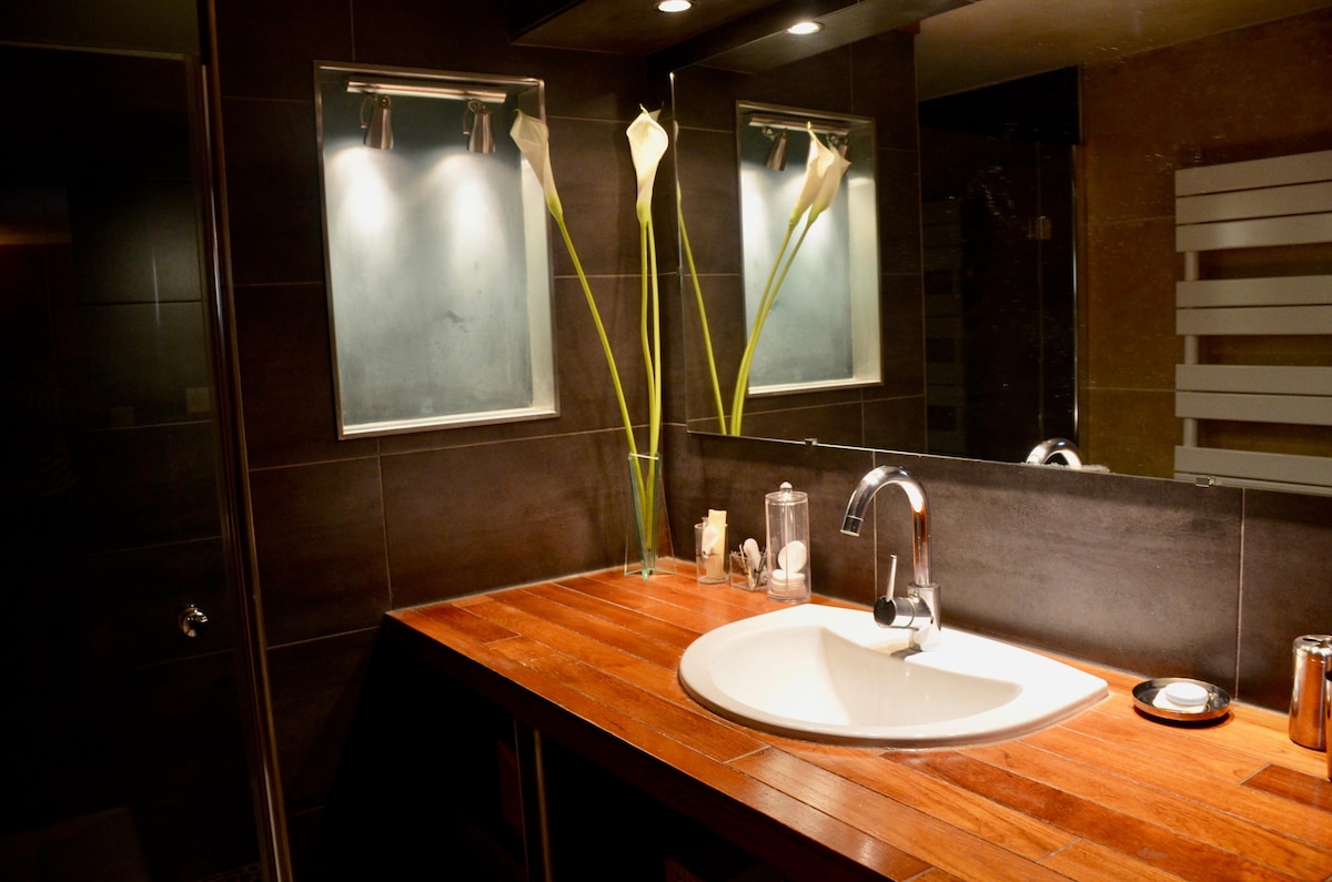A modern bathroom is presented with a polished wooden countertop and a circular sink. Tall, slender flower vases with white flowers are positioned near the mirror, which is surrounded by soft lighting. A glass shower enclosure and various toiletries are visible in the space.