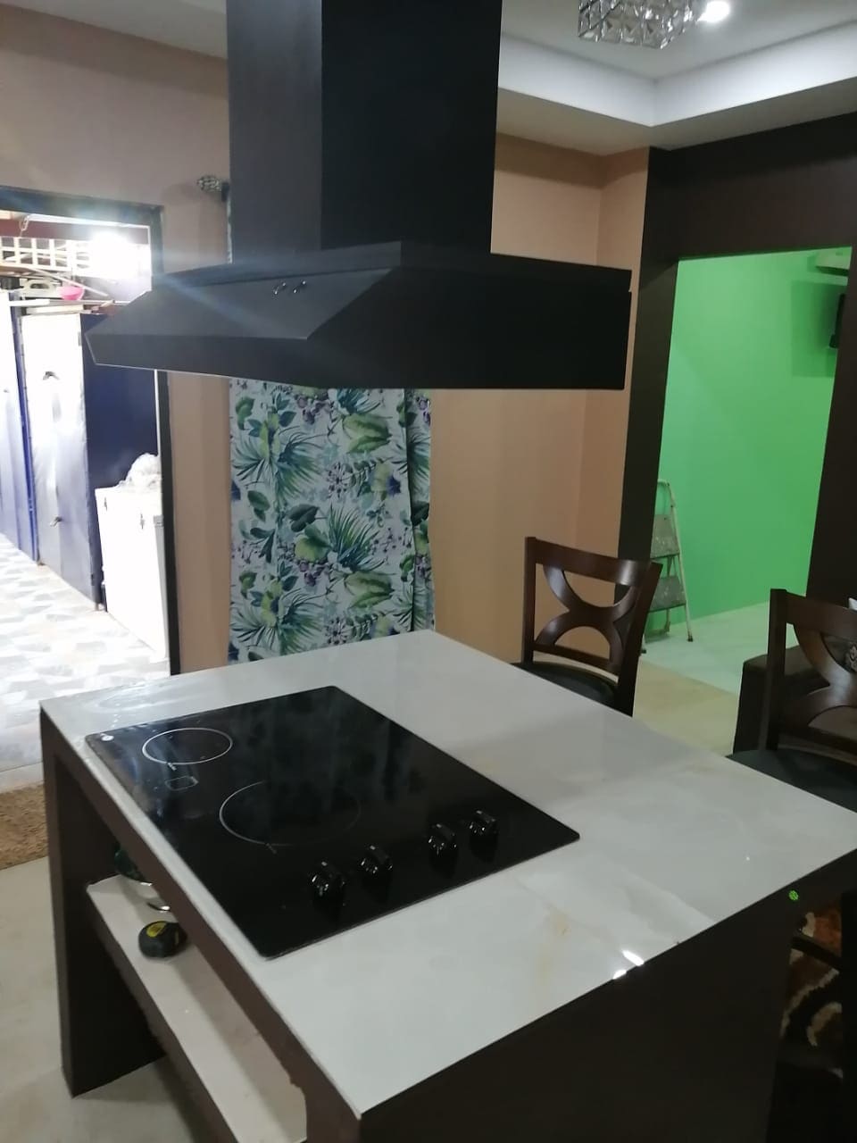 A modern kitchen area is highlighted by a sleek cooking island with a built-in cooktop. Dark cabinetry complements the marble countertop, while stylish chairs are positioned around the island. A vibrant curtain adds a touch of color, and additional living spaces are visible in the background.