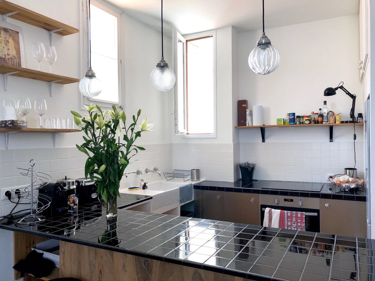 A modern kitchen is presented with sleek black tiled countertops and light wooden shelving. Fresh lilies are arranged in a vase at the center. Natural light enters through two windows, illuminating the space. Essential appliances and pantry items are neatly organized along the walls.