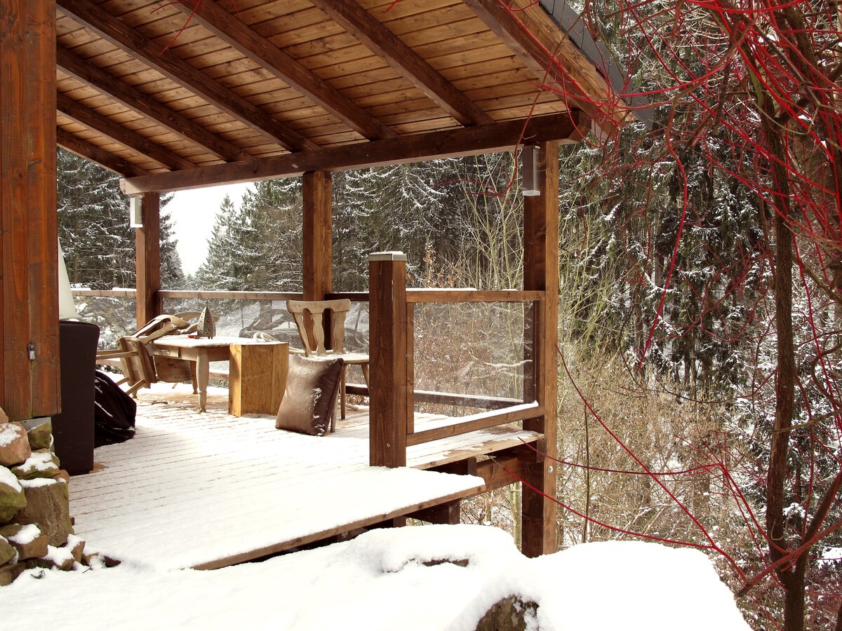 A covered wooden terrace is seen, featuring a rustic table and several chairs made of natural materials. Snow blankets the floor and surrounding area, while trees are visible in the background, providing a serene winter atmosphere.